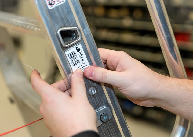 Fachkraft der i-Arbeitsschutz bringt nach einer erfolgreichen Betriebsmittelprüfung eine Prüfplakette mit Barcode an einer Aluminiumleiter an.