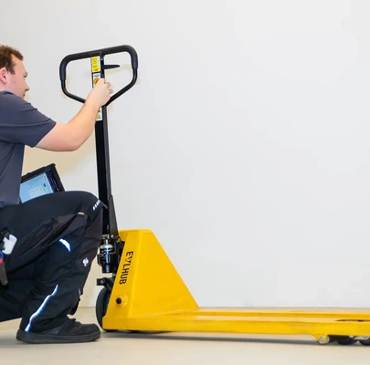 Prüfer der i-Arbeitsschutz Service GmbH kontrolliert die Mechanik eines gelben Handhubwagens im Rahmen der jährlichen UVV-Prüfung für Flurförderzeuge.
