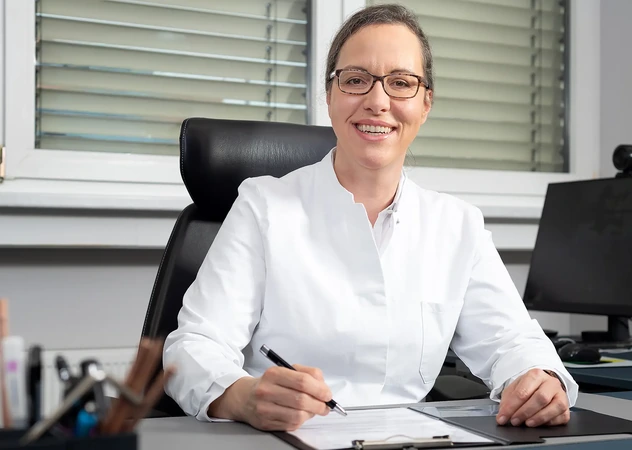 Freundliche, erfahrene Betriebsärztin der i-Arbeitsschutz Service GmbH lächelt am Schreibtisch in ihrem Behandlungsraum.